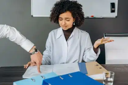 Zahnarztpraxis-Mitarbeiterin am Schreibtisch mit Papierstapeln und Bildschirmen, angespannte Körperhaltung